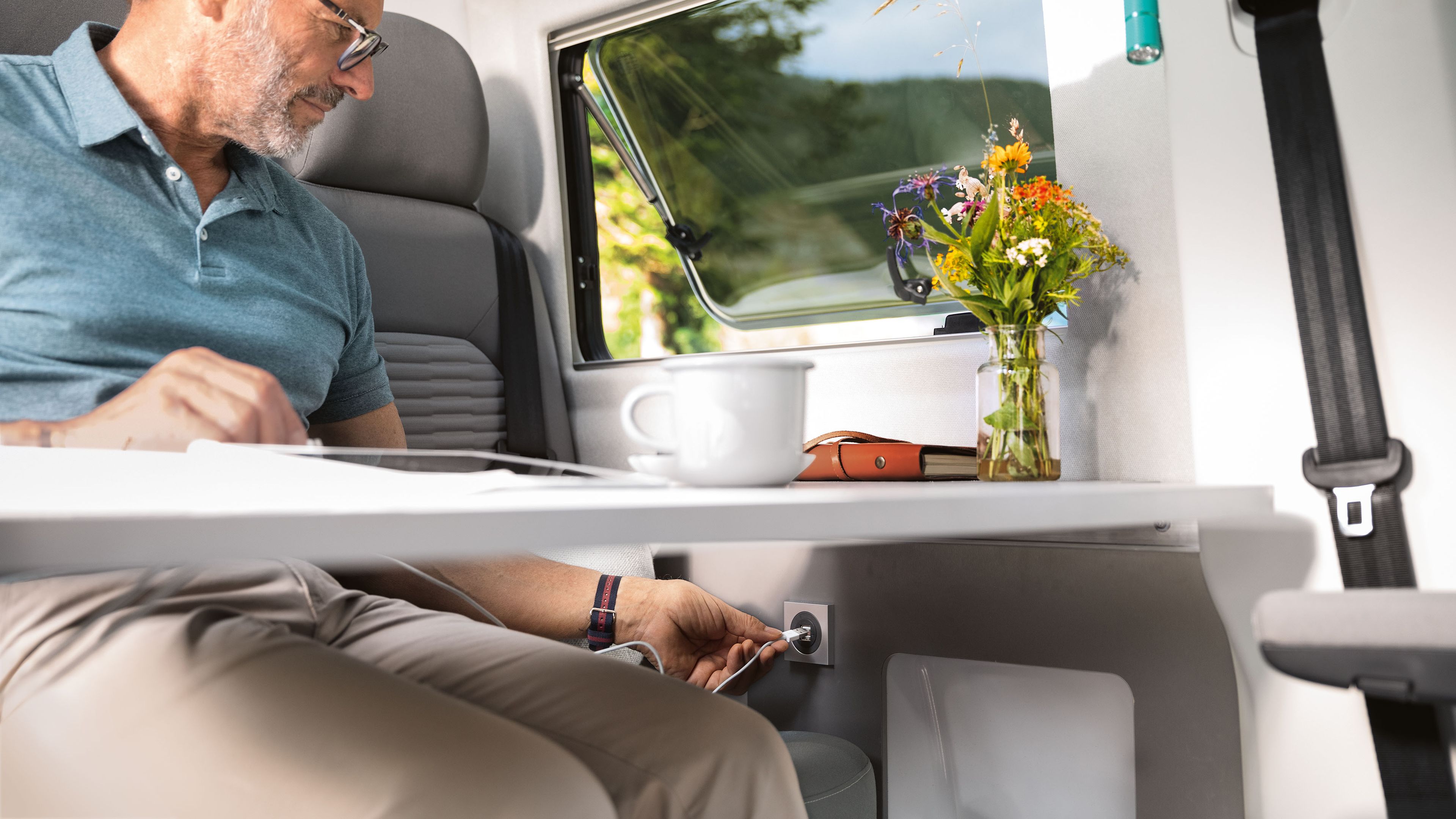 Um homem está sentado à mesa e está a ligar o cabo de carregamento do seu tablet à tomada na parede do veículo. 