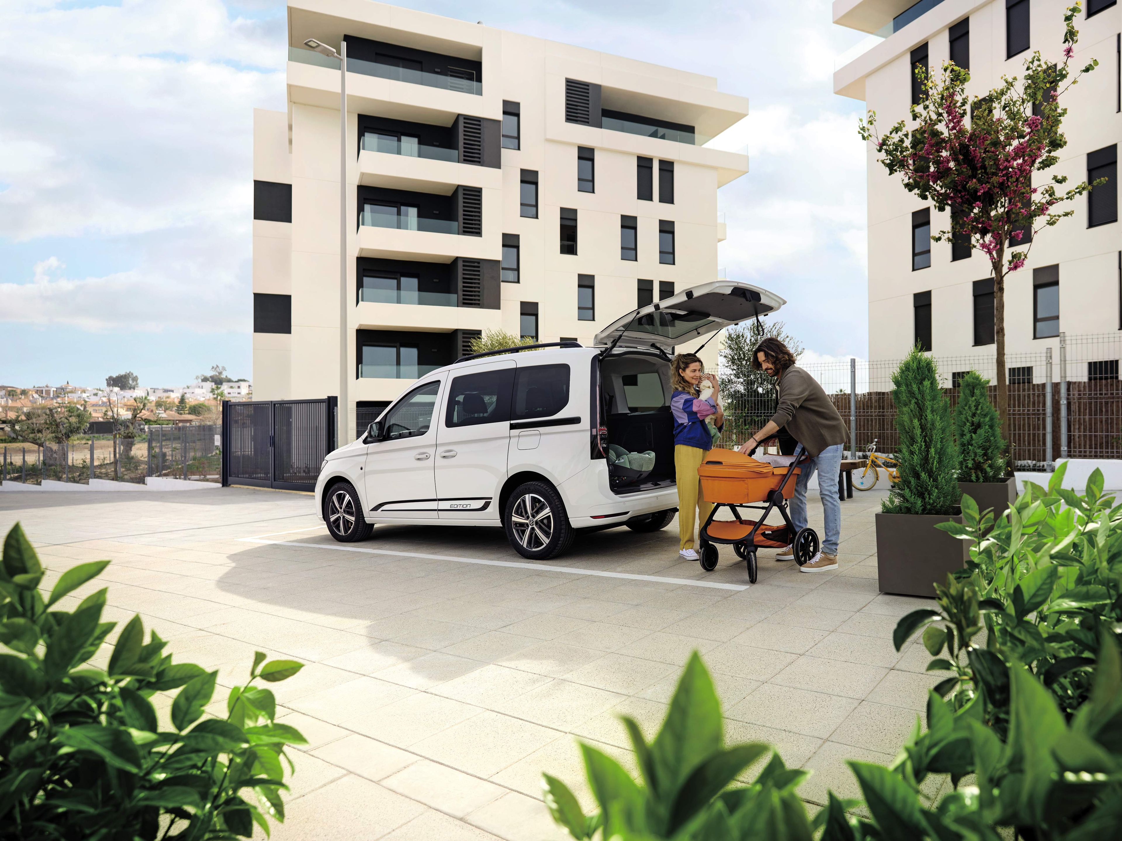 Eltern verstauen einen Kinderwagen im Kofferraum eines VW Caddy, Mutter hält das Baby
