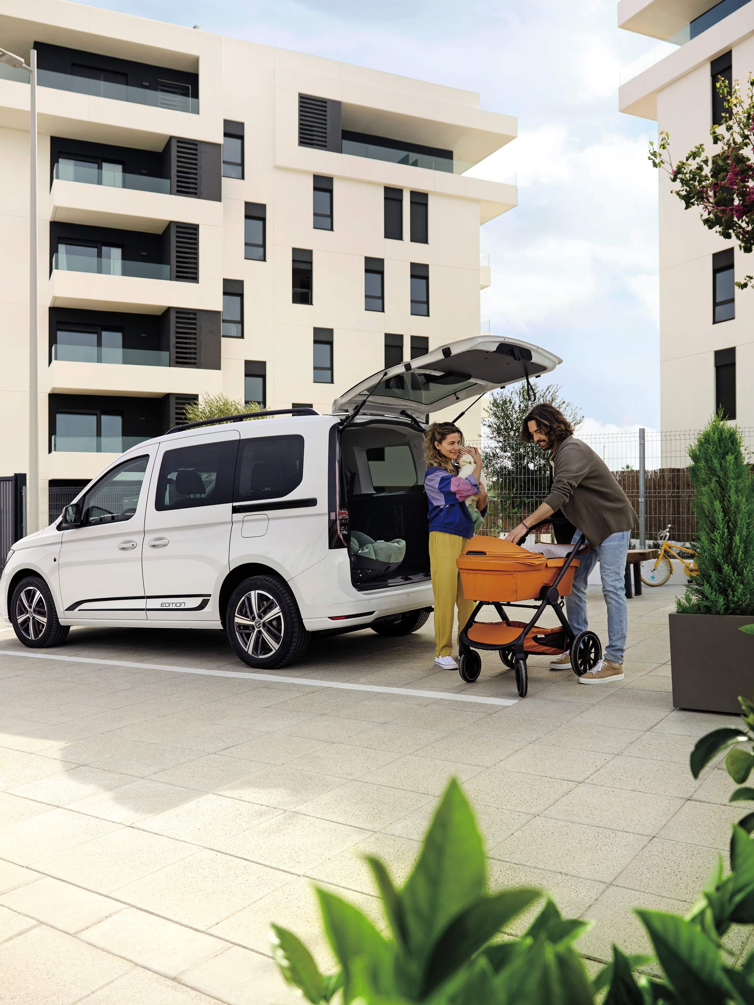 Eltern verstauen einen Kinderwagen im Kofferraum eines VW Caddy, Mutter hält das Baby