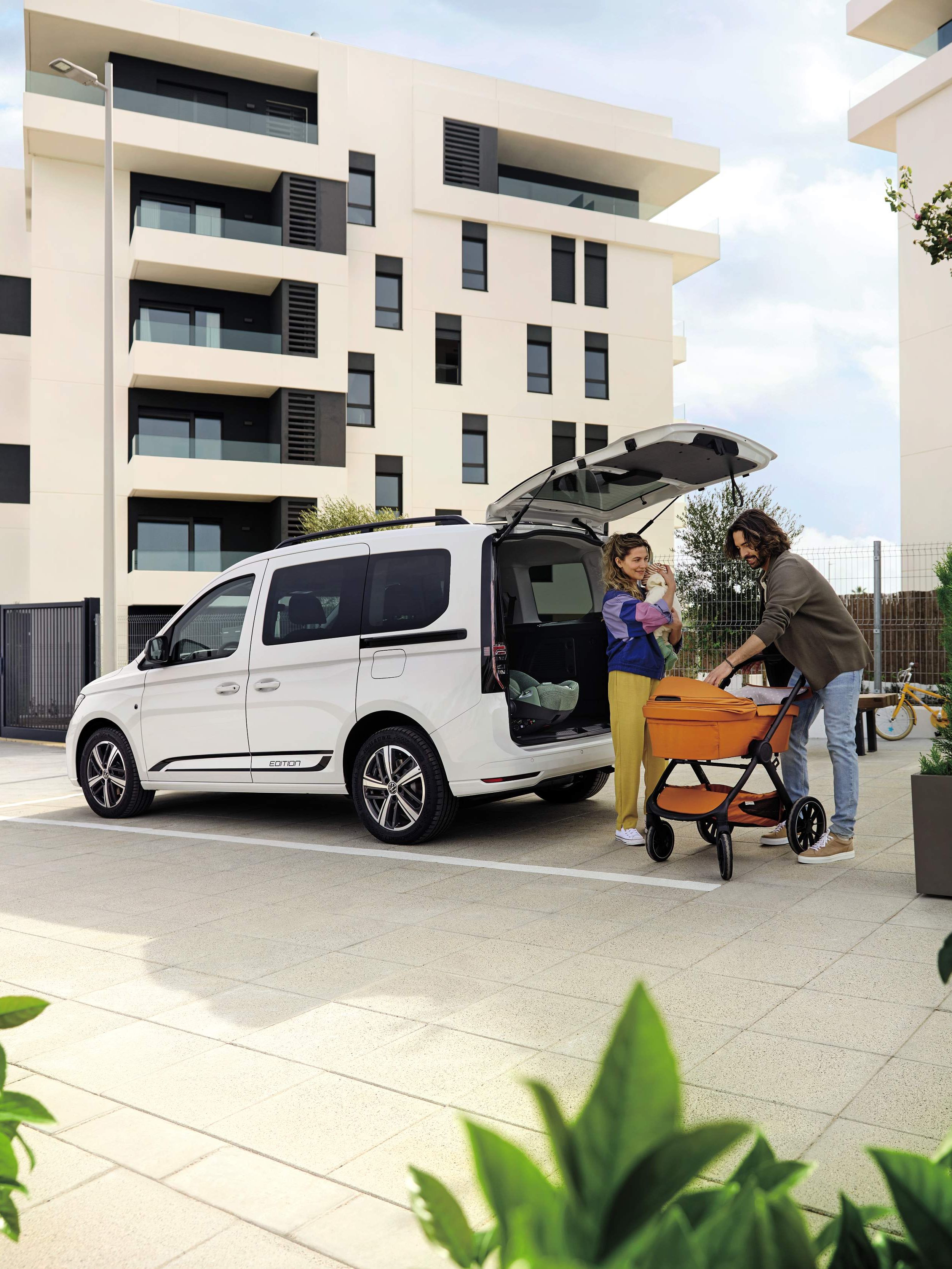 Eltern verstauen einen Kinderwagen im Kofferraum eines VW Caddy, Mutter hält das Baby