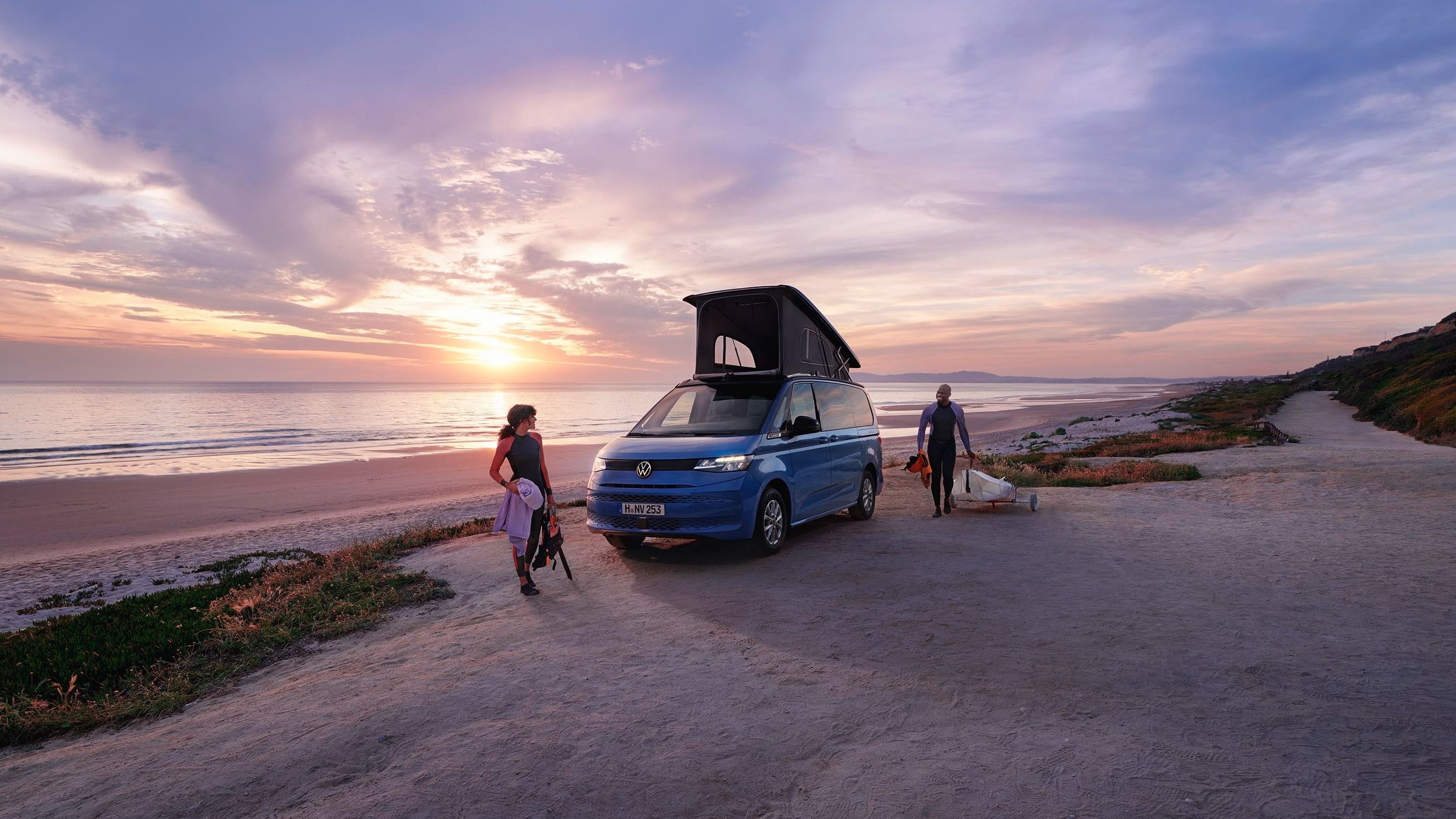 Dois surfistas na praia ao amanhecer com uma Volkswagen California.