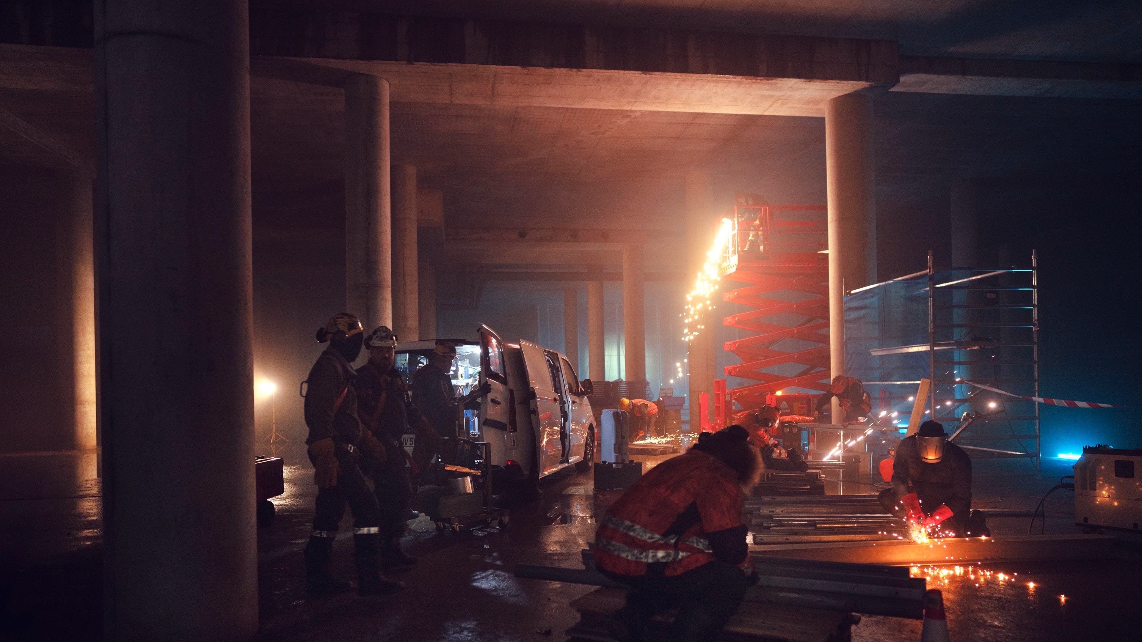 Trabalhadores da construção civil cortam metal, faúlhas iluminam a cena.