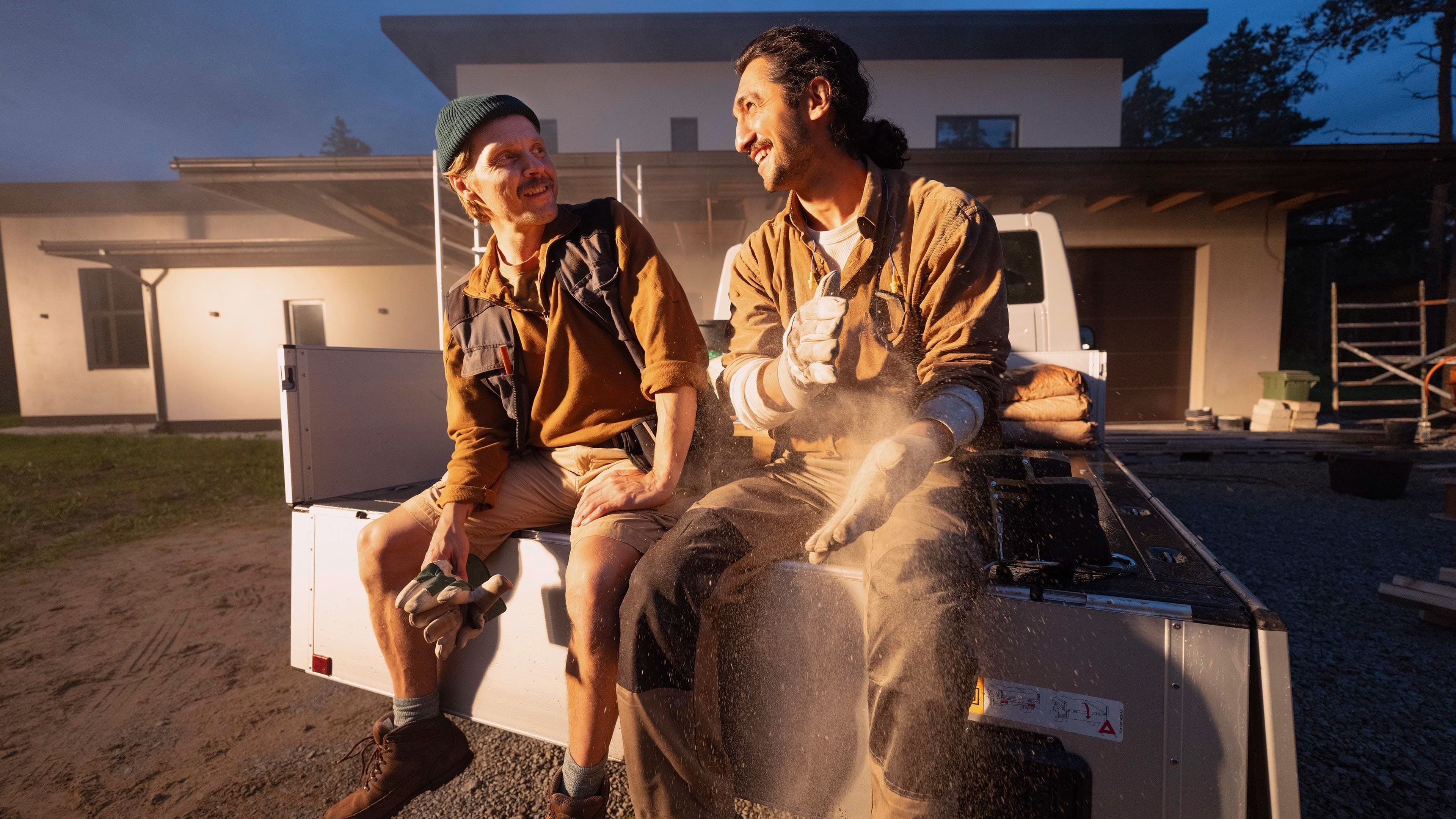 Dois trabalhadores da construção civil sentados na traseira de um Volkswagen, há poeira no ar.