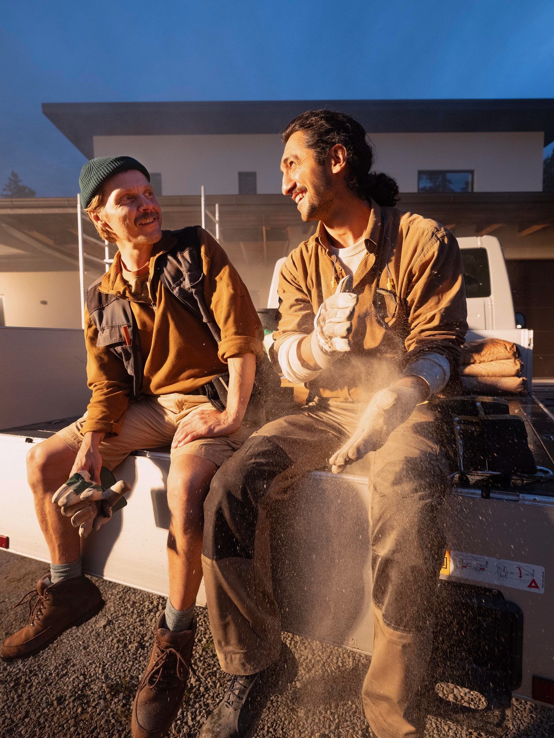 Dois trabalhadores da construção civil sentados na traseira de um Volkswagen, há poeira no ar.