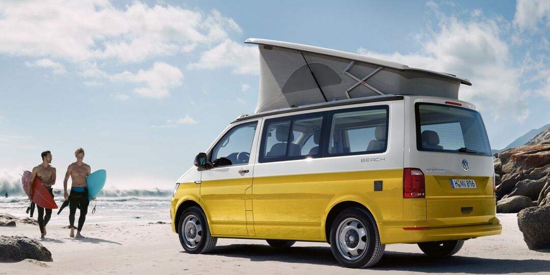 Uma California da Volkswagen Veículos Comerciais amarela, na praia, iluminada pelo sol. Dois surfistas a correr na sua direção.