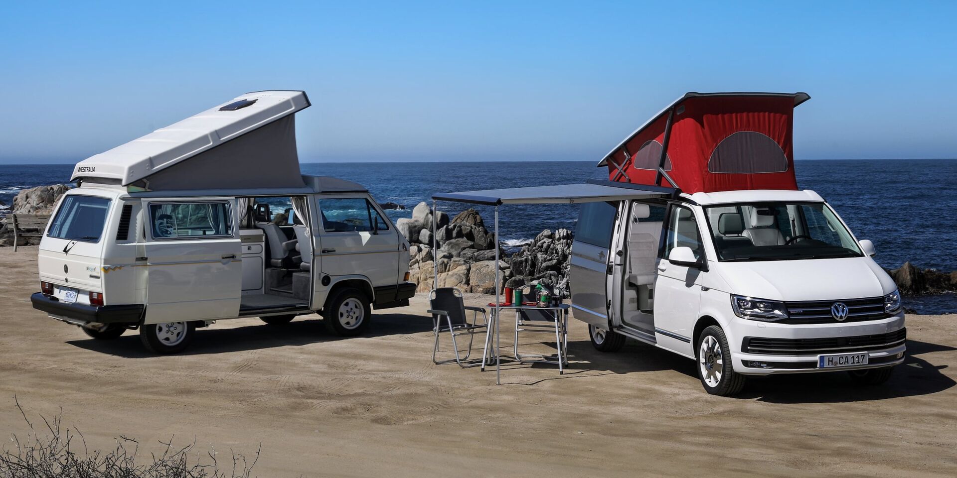 Duas California da Volkswagen Veículos Comerciais com tejadilho elevado numa praia escarpada. O toldo do veículo à direita está estendido. Algum do equipamento de campismo encontra-se sob este.