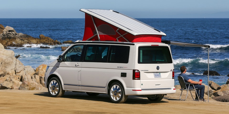 Uma California da Volkswagen Veículos Comerciais com tejadilho elevado vermelho, na praia. O toldo está estendido. O condutor observa o mar.
