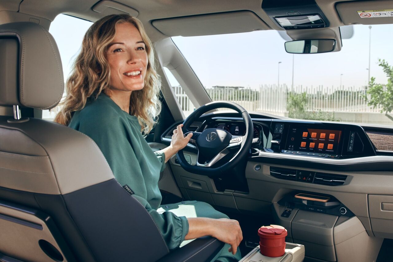 Uma vista do habitáculo a partir da fila de bancos traseiros num VW Multivan. Uma mulher está sentada ao volante.