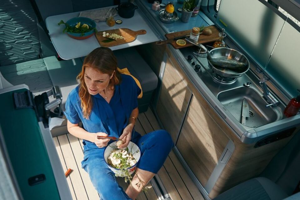 Uma mulher está sentada no chão da cozinha da autocaravana California 6.1 a preparar uma salada.