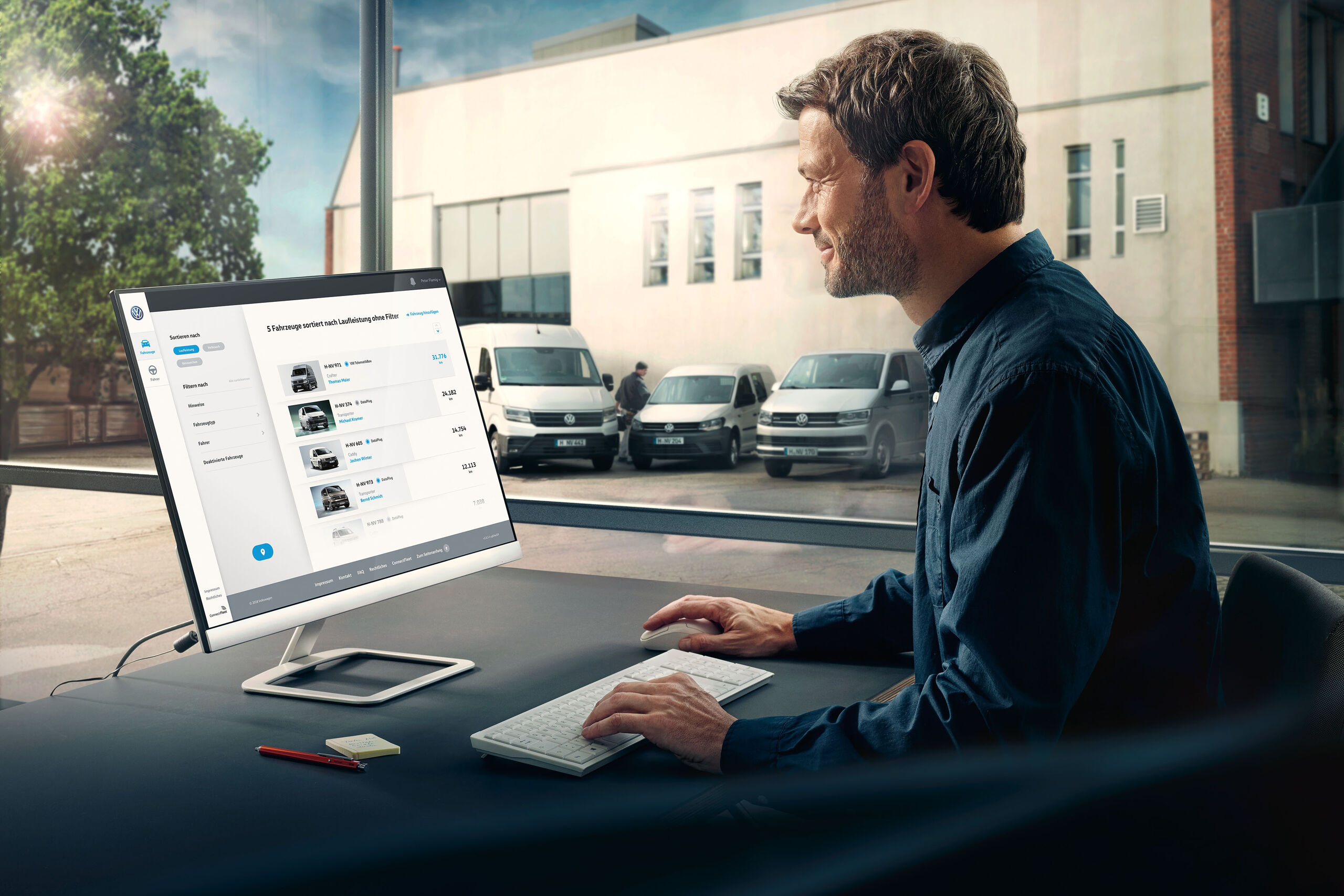 Um homem está sentado no escritório com um computador à frente. No ecrã pode ver-se o sistema de gestão de frotas We Connect Fleet da Volkswagen. Através da janela, pode ver-se o estacionamento da empresa com três carrinhas da Volkswagen Veículos Comerciais.