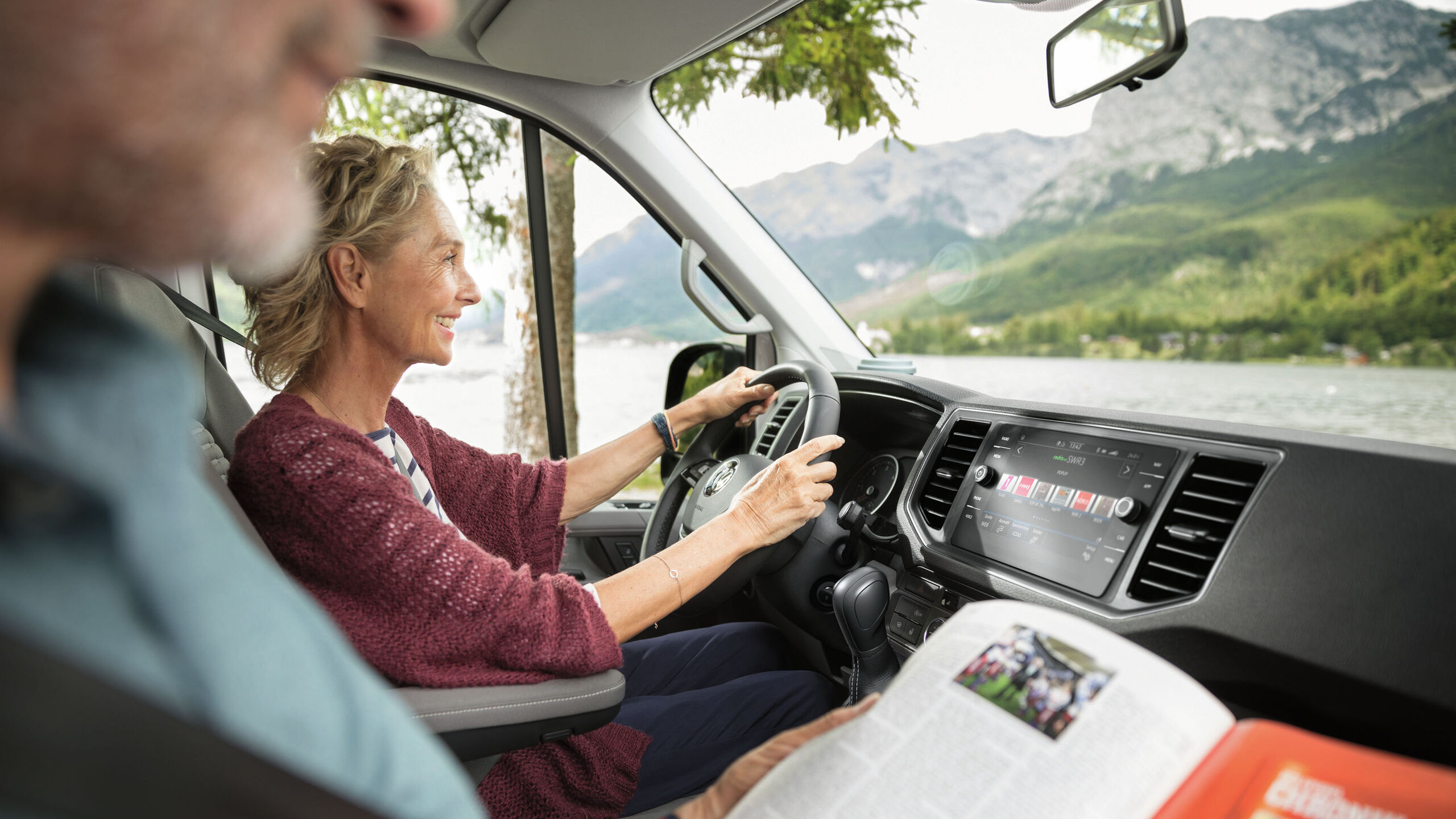 Mulher sentada ao volante do VW Grand California, homem sentado ao lado dela a ler um jornal.