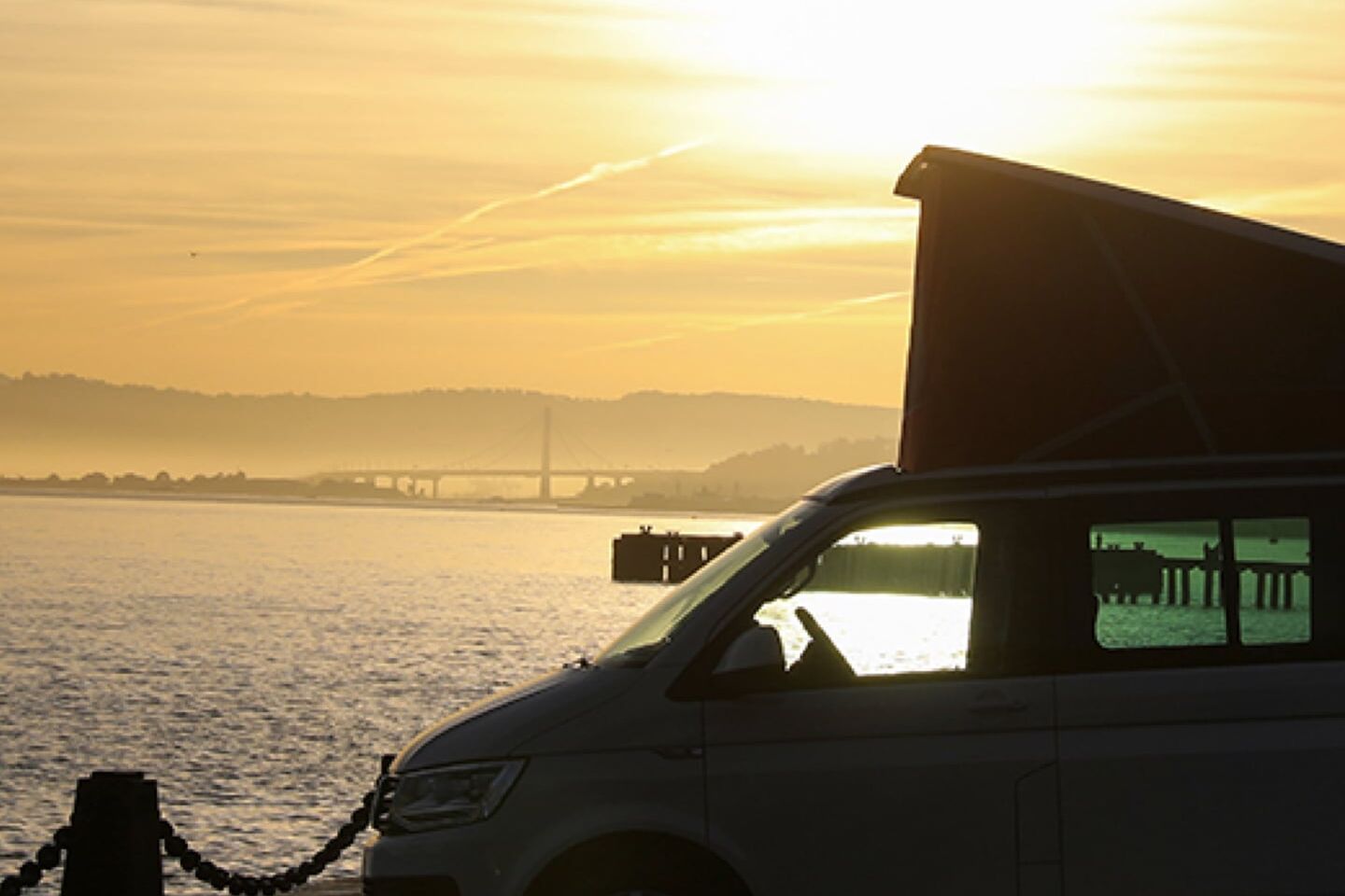 A California da Volkswagen Veículos Comerciais em contra-luz ao pôr do sol numa estrada costeira à beira-mar.