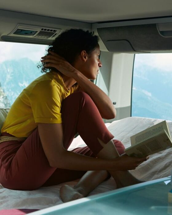 Uma mulher sentada na cama da VW California 6.1 a olhar para a paisagem nas montanhas.