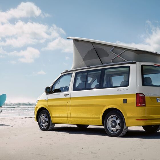 Uma California da Volkswagen Veículos Comerciais amarela, na praia, iluminada pelo sol. Dois surfistas a correr na sua direção.