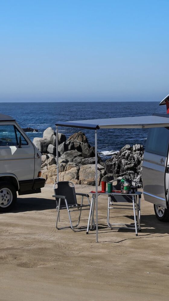 Duas California da Volkswagen Veículos Comerciais com tejadilho elevado numa praia escarpada. O toldo do veículo à direita está estendido. Algum do equipamento de campismo encontra-se sob este.