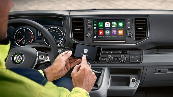 Na cabina do condutor da carrinha VW Crafter parada. No fundo pode ver-se o serviço online Car-Net no ecrã multifunções. Em primeiro plano, um homem mexe no seu smartphone.