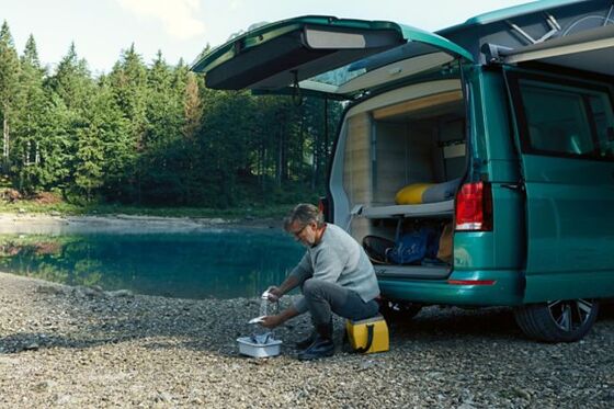 VW California 6.1 steht vor einem Wald. Ein Mann nutzt die Außendusche zum Geschirrspülen.