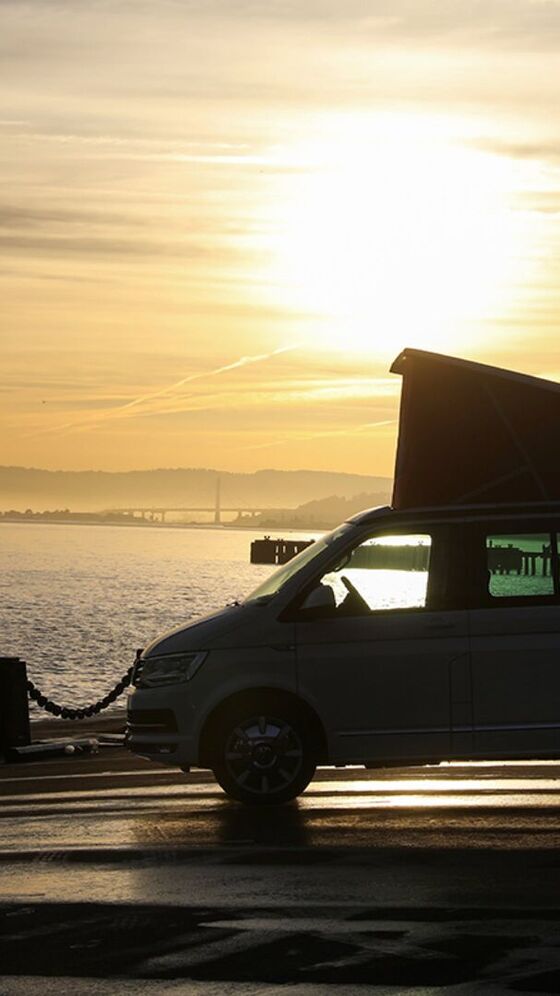 A California da Volkswagen Veículos Comerciais em contra-luz ao pôr do sol numa estrada costeira à beira-mar.