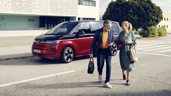Um homem e uma mulher caminham em frente a uma Multivan VW.